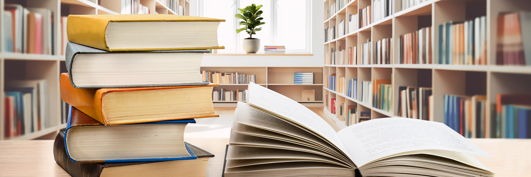 An open book and a stack of closed books sit on a table in front of wall-to-wall bookshelves.