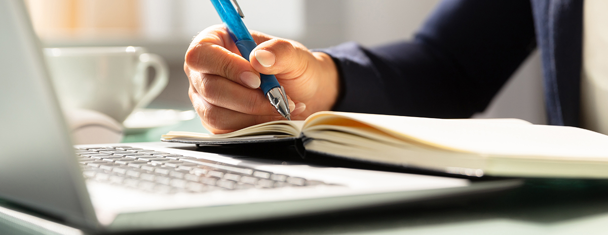 A person writing in a notebook while sitting in front of a laptop, with a coffee cup in the background.